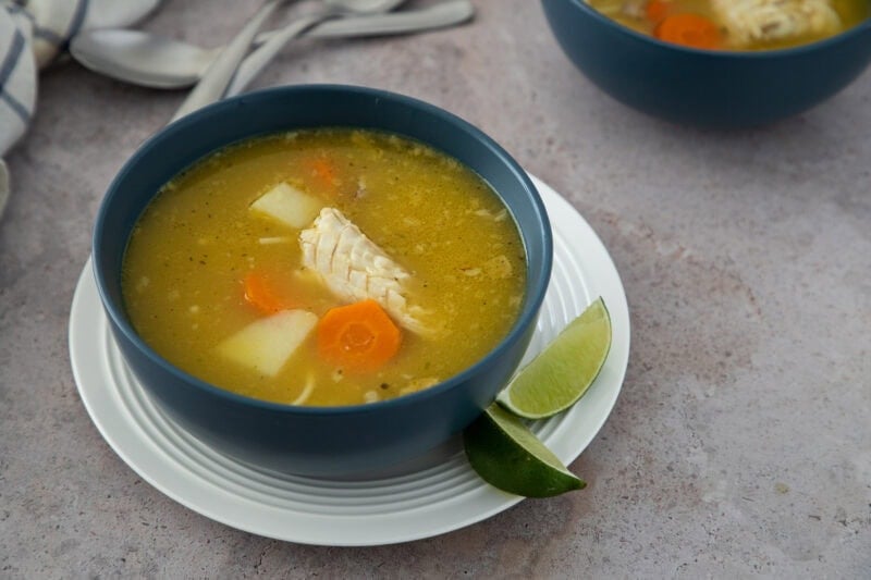 Dominican chicken soup with yuca, carrots, and tender chicken, served in a blue bowl with lime wedges.