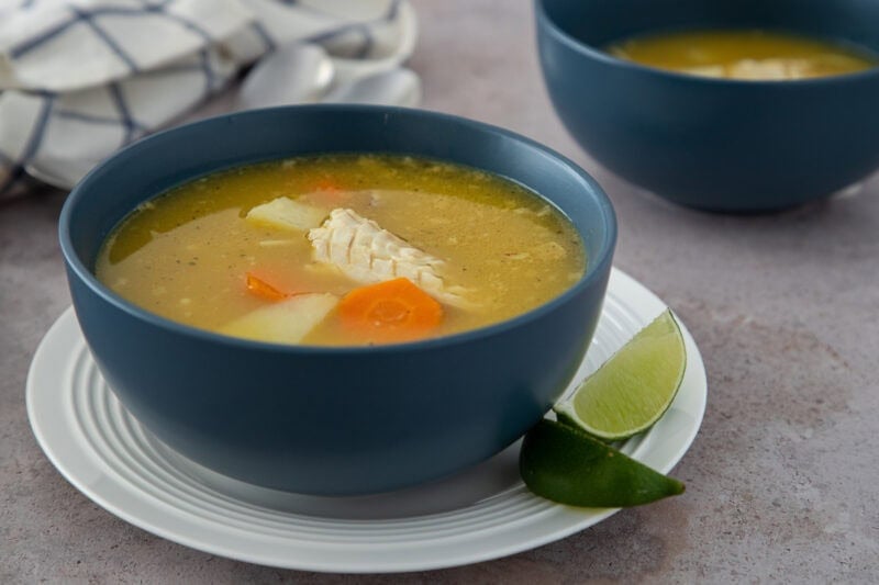 Dominican chicken soup with yuca, carrots, and tender chicken, served in a blue bowl with lime wedges.