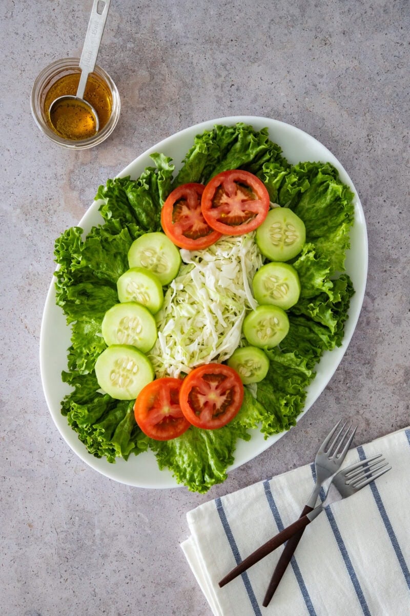 Close-up of a green salad with lettuce, cucumbers, tomatoes, and cabbage on a plate, with dressing and forks nearby.