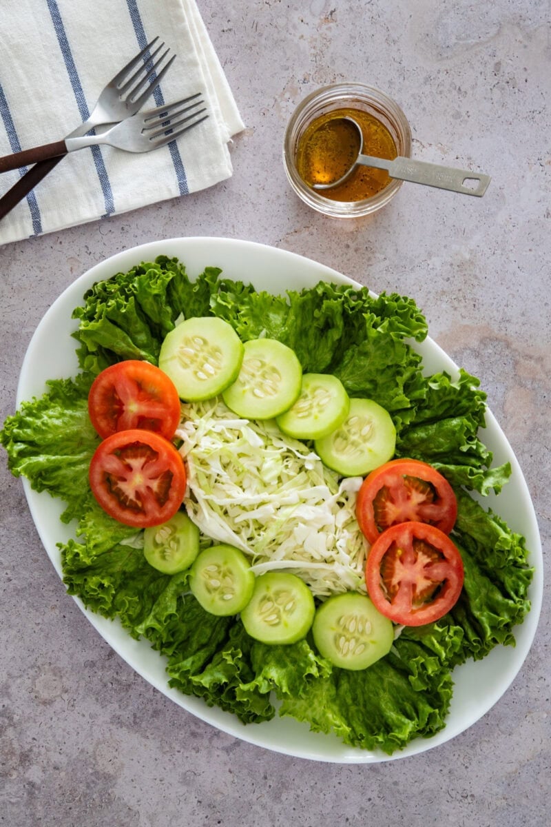 Overhead view of a fresh salad with lettuce, cucumber slices, tomato slices, and shredded cabbage, alongside a small bowl of dressing.