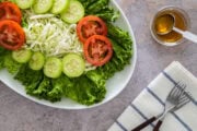 Fresh green salad with lettuce, sliced cucumbers, tomatoes, and shredded cabbage on a white plate, with dressing on the side.